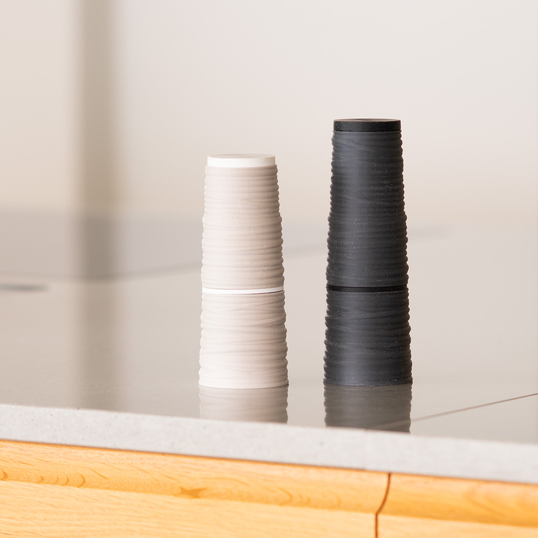 Two conic textured salt & pepper mills, one white and one black, on a reflective surface with a blurred background.