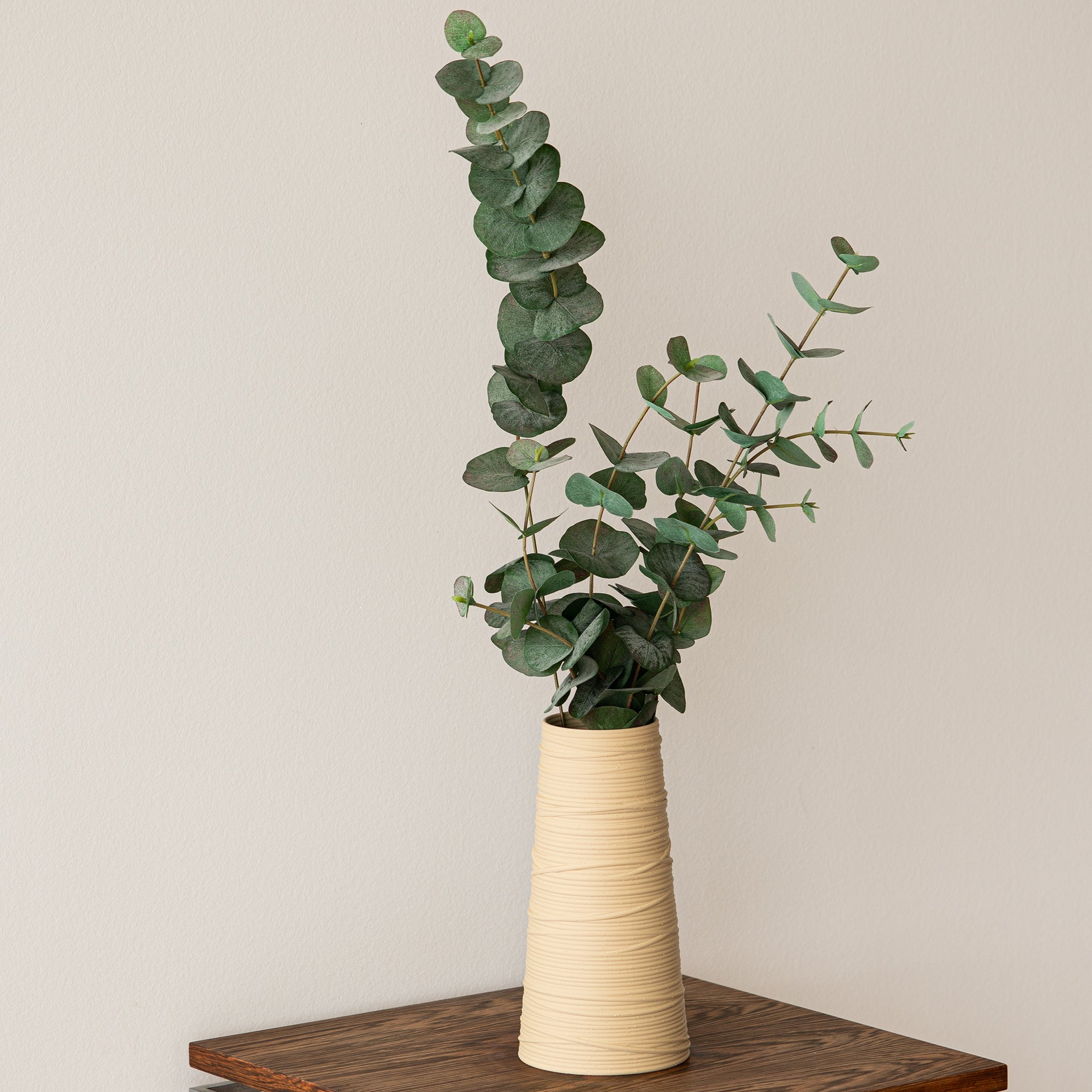 Beige textured vase with green eucalyptus on a wooden surface against a beige wall