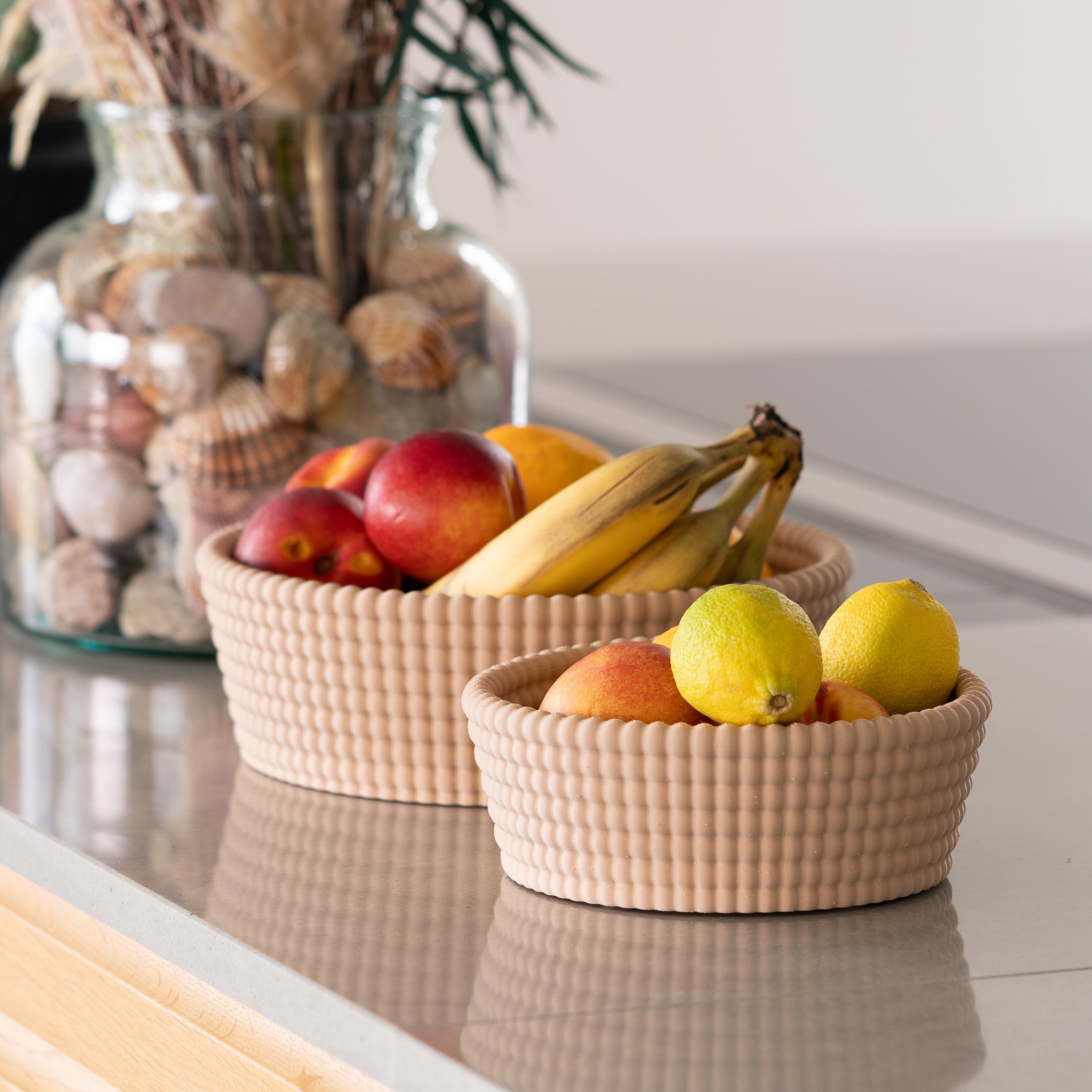 Three woven baskets on a countertop with fruits and a decorative jar in the background.