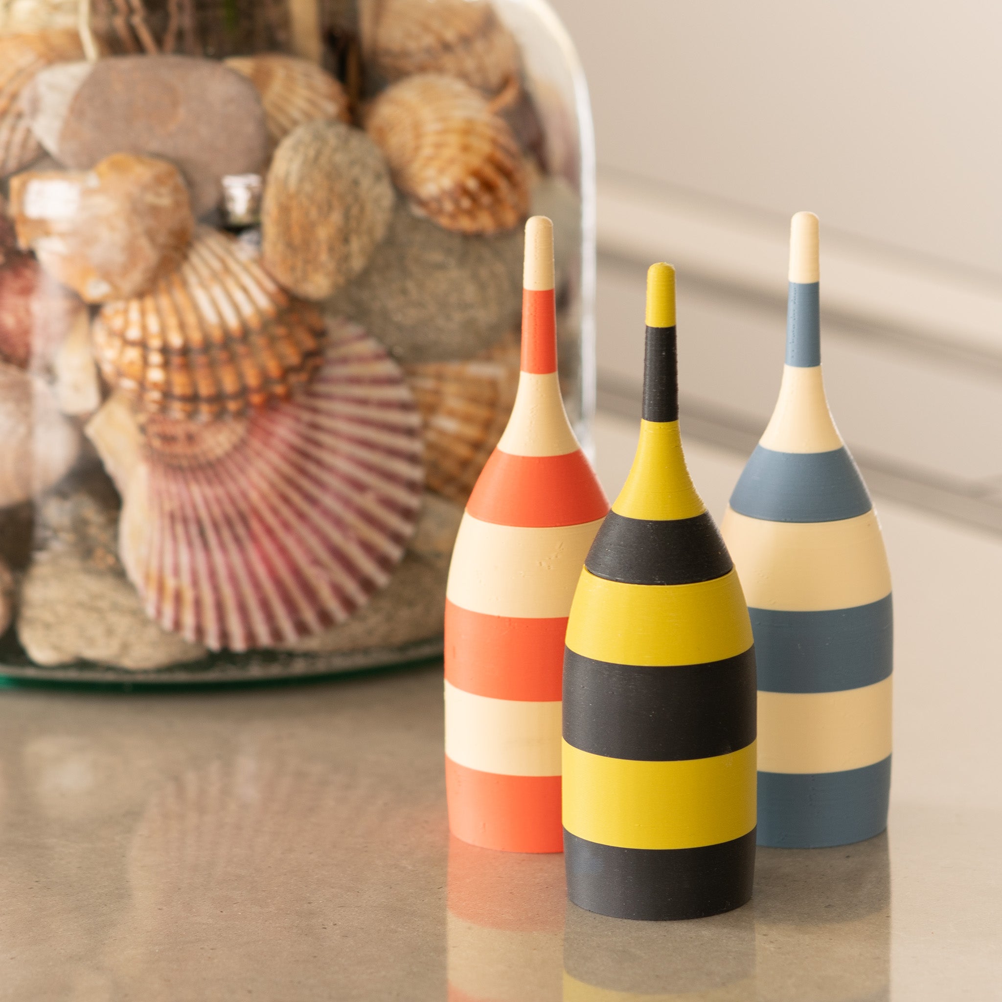 Three colorful striped toothpick dispensers on a reflective surface with a jar of seashells in the background.
